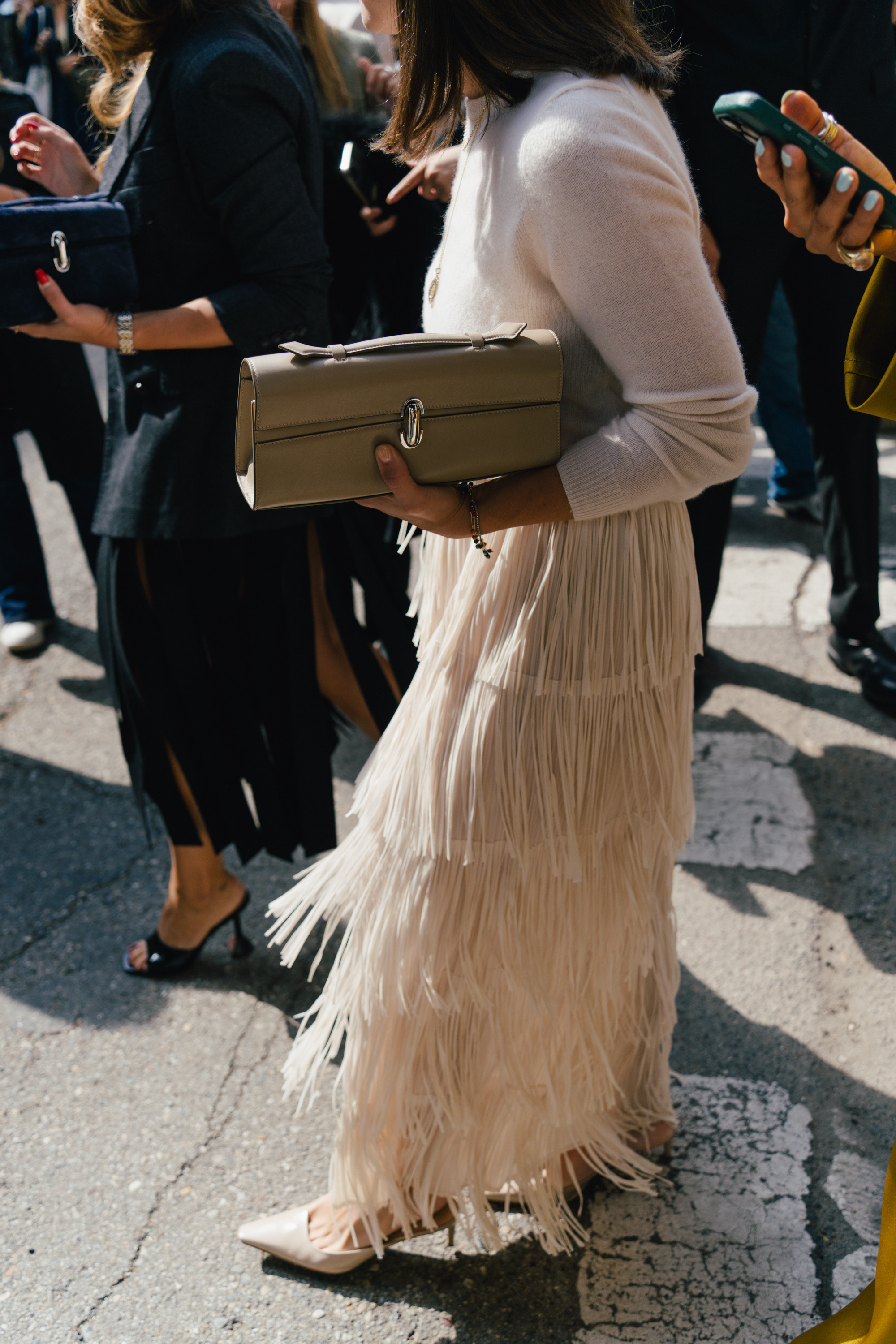 A street style photo of a person in a cream fringed outfit holding a structured beige clutch while standing among a crowd on a sunny city street.
