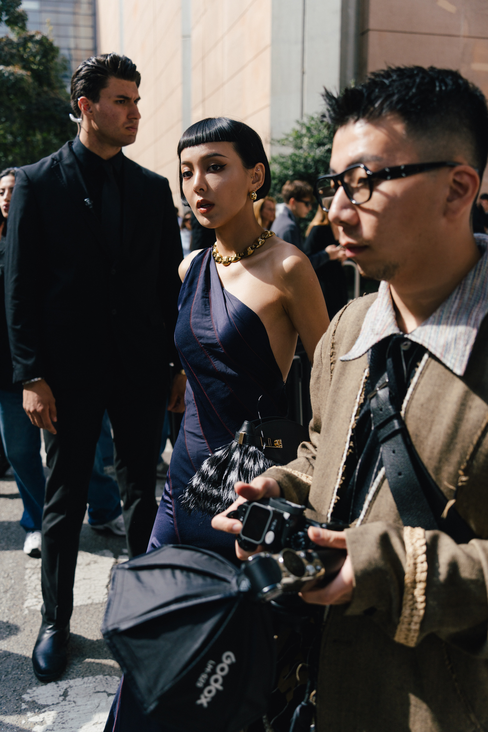 A street style photo of a person in a one-shoulder dark dress with a gold necklace, standing in a crowd beside photographers and security staff.