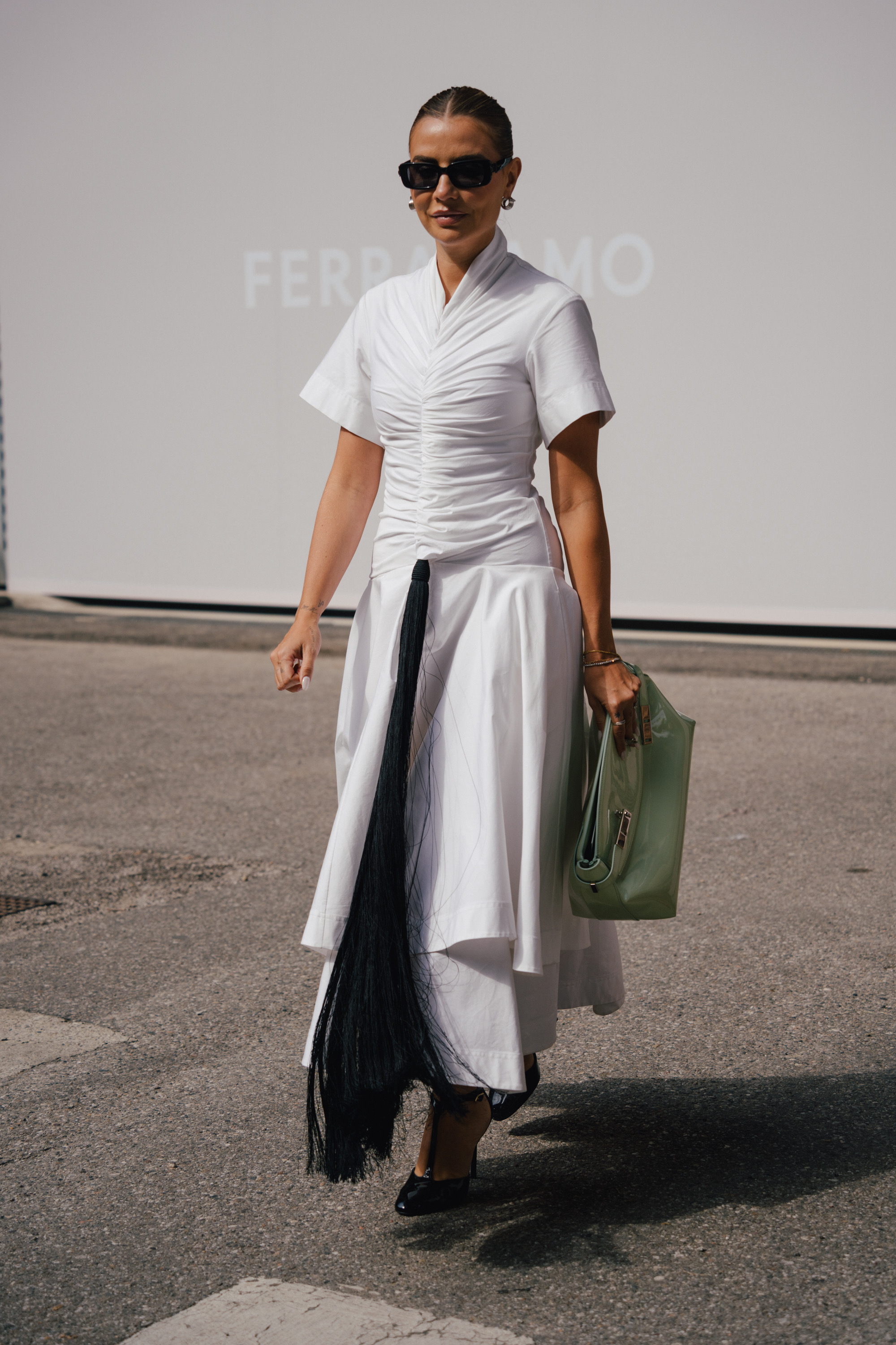 A street style photo of a person wearing sunglasses and a white ruched dress with a long black tassel detail, walking while carrying a translucent green handbag.