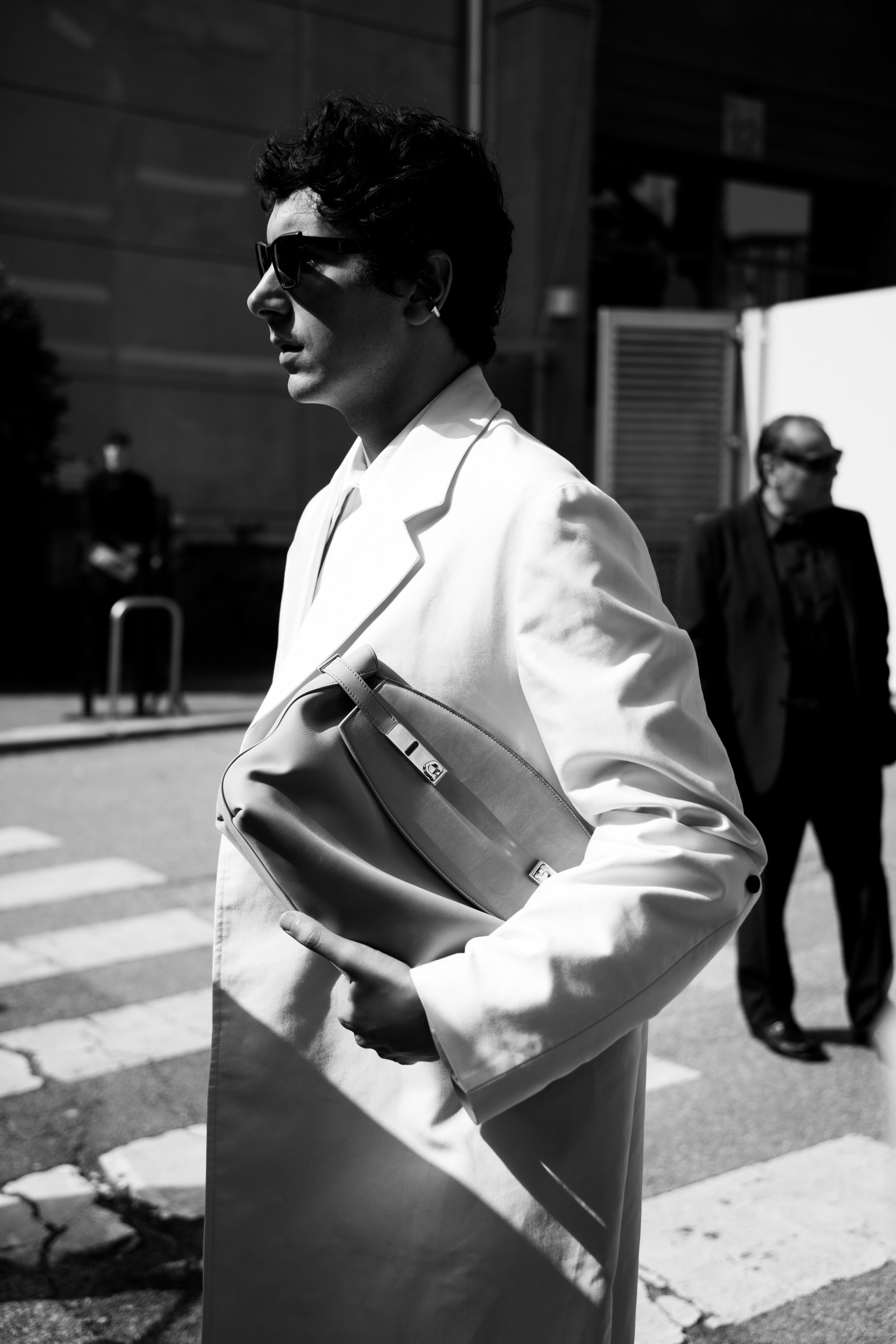 A black-and-white street style photo of a person wearing sunglasses and a light-colored blazer, holding a large clutch while walking across a city crosswalk.