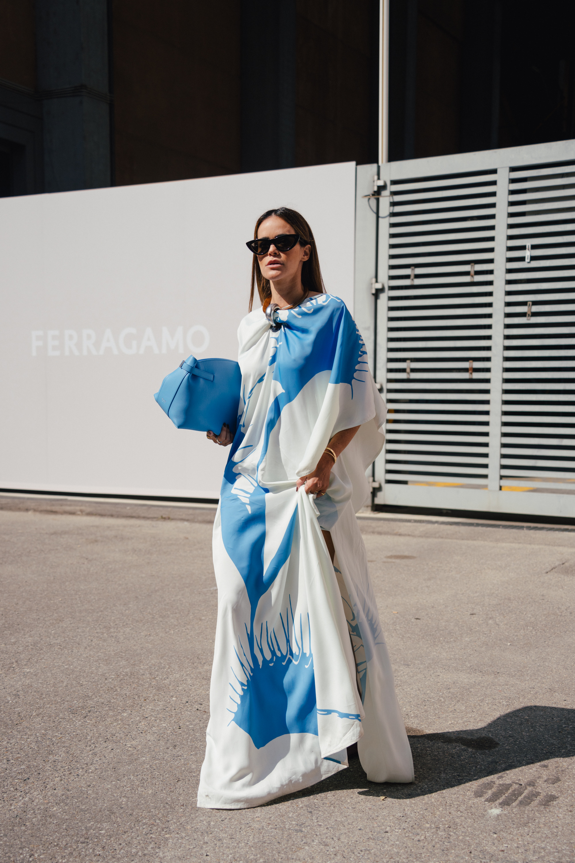 A street style photo of a person wearing sunglasses and a flowing white outfit with bold blue abstract patterns, walking while carrying a matching blue clutch.