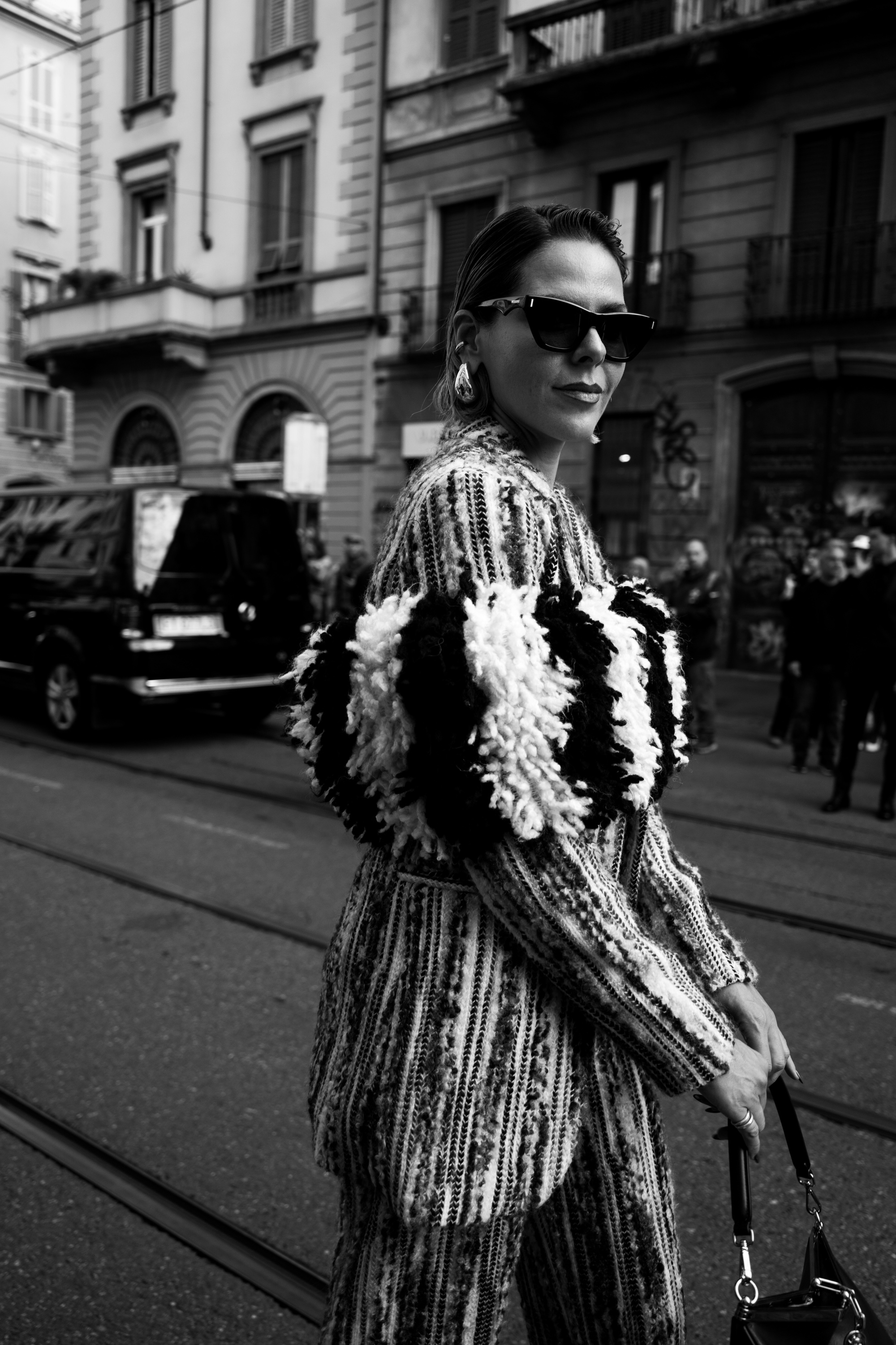 A black-and-white street style photo of a person wearing sunglasses and a textured patterned outfit with bold black-and-white fuzzy sleeve details, standing on a city street with a handbag.