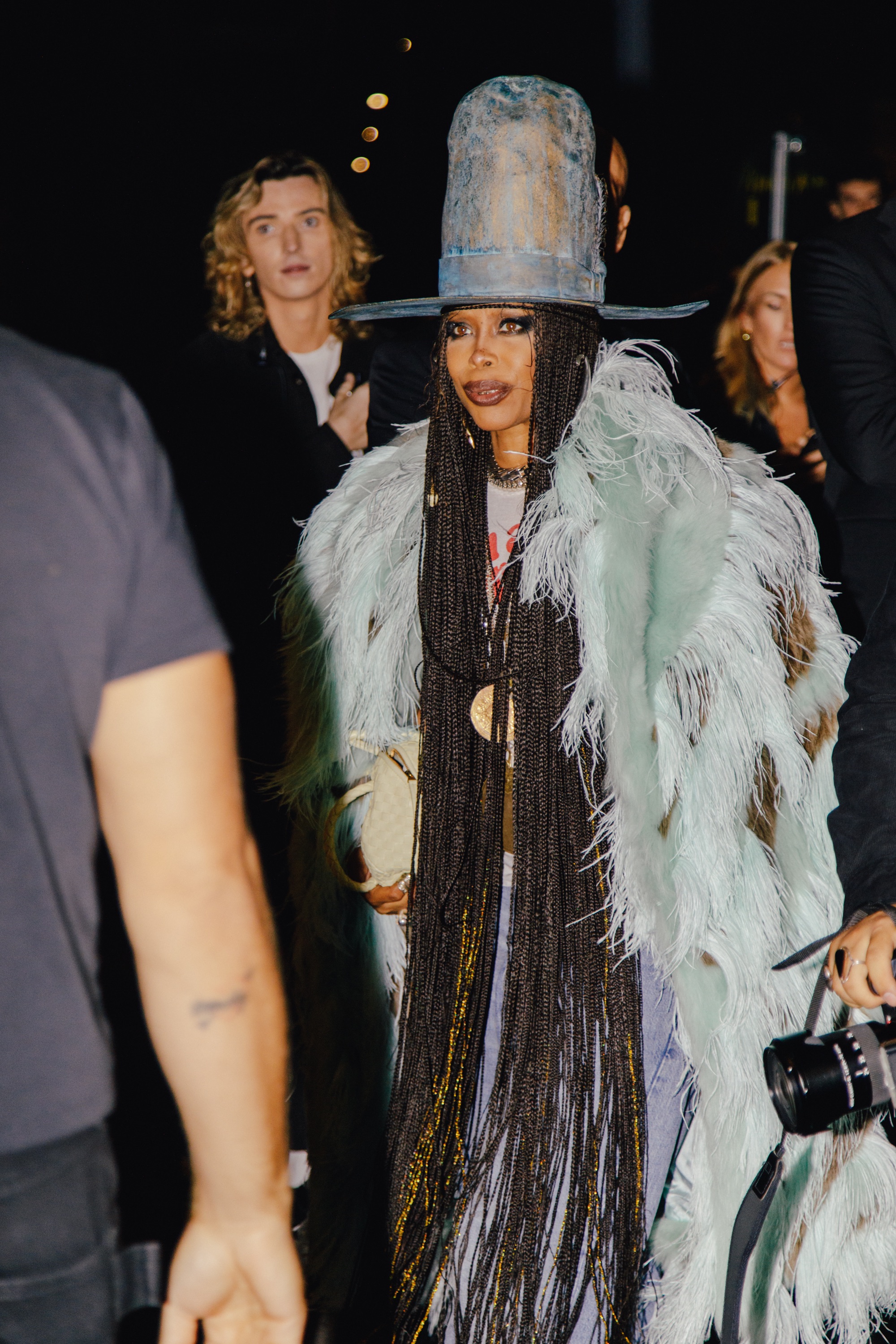 A street style photo of a person wearing a tall blue hat, long braids, and a pale blue feathered coat, standing in a nighttime crowd.