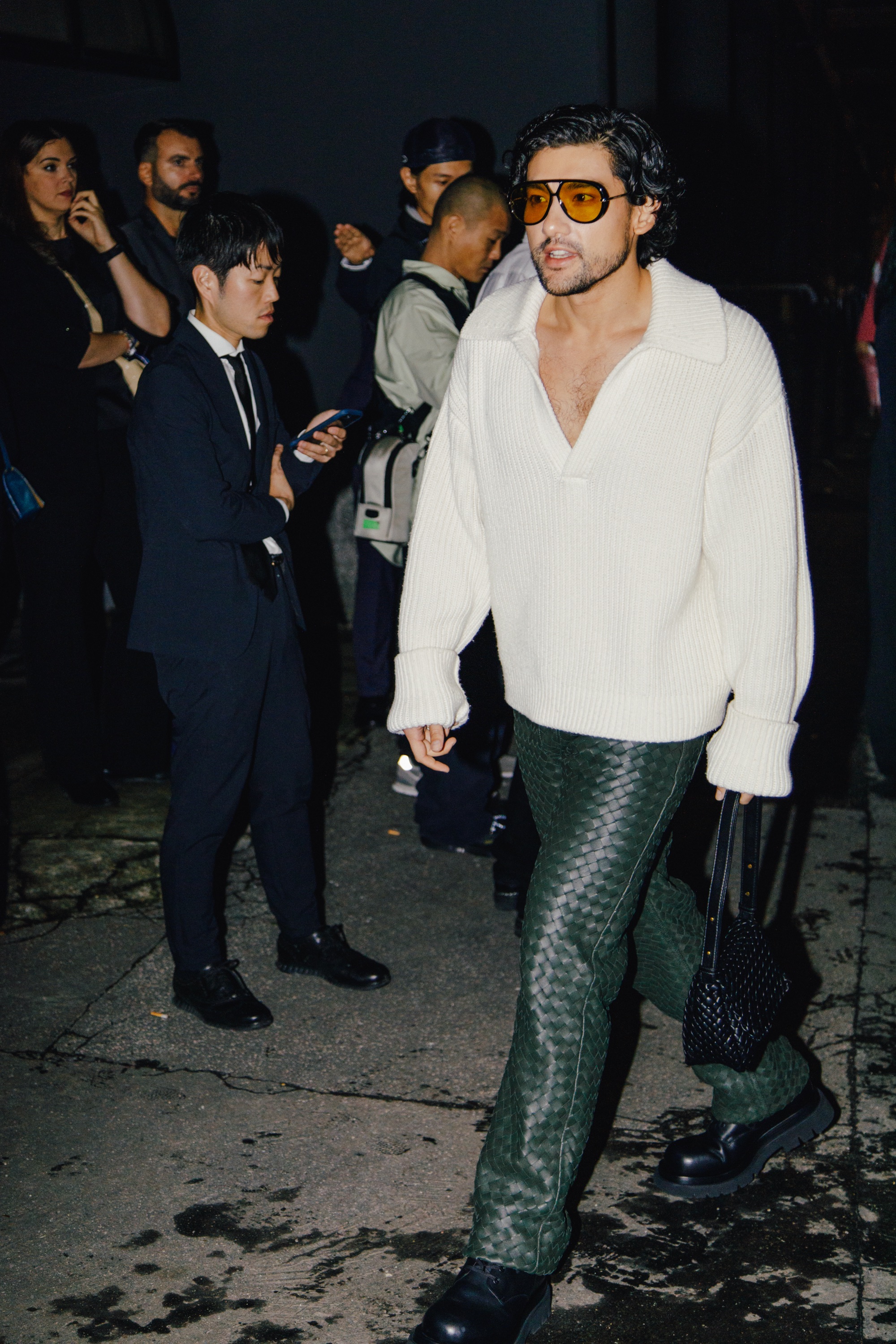 Man wearing amber-tinted sunglasses, an oversized cream knit sweater, green woven-texture trousers, and black shoes, carrying a black woven bag while walking past people at night.