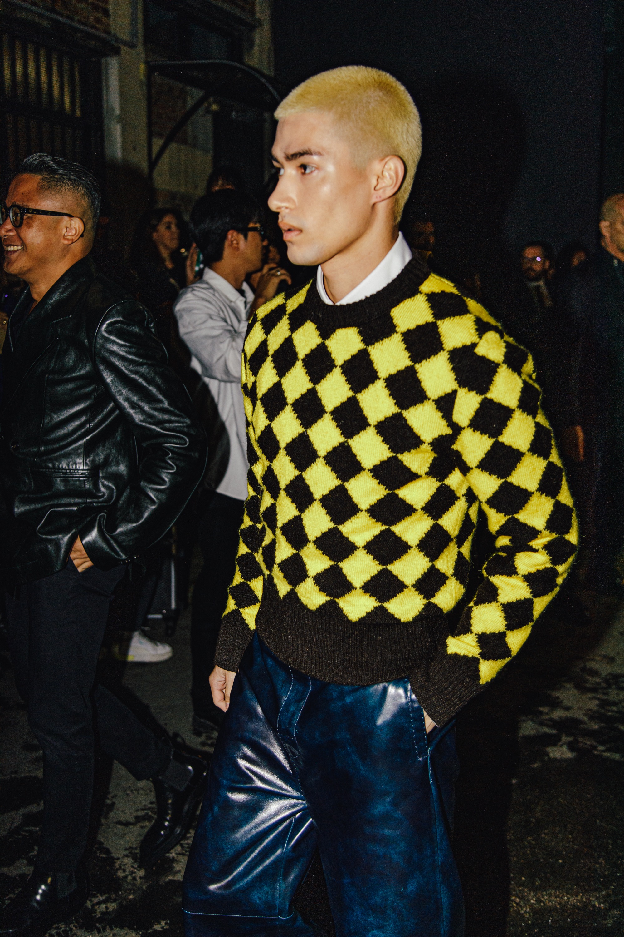 Young man with bleached blond hair wearing a fuzzy yellow-and-black diamond-patterned sweater over a white collared shirt, paired with glossy dark blue trousers, walking with one hand in his pocket at night.