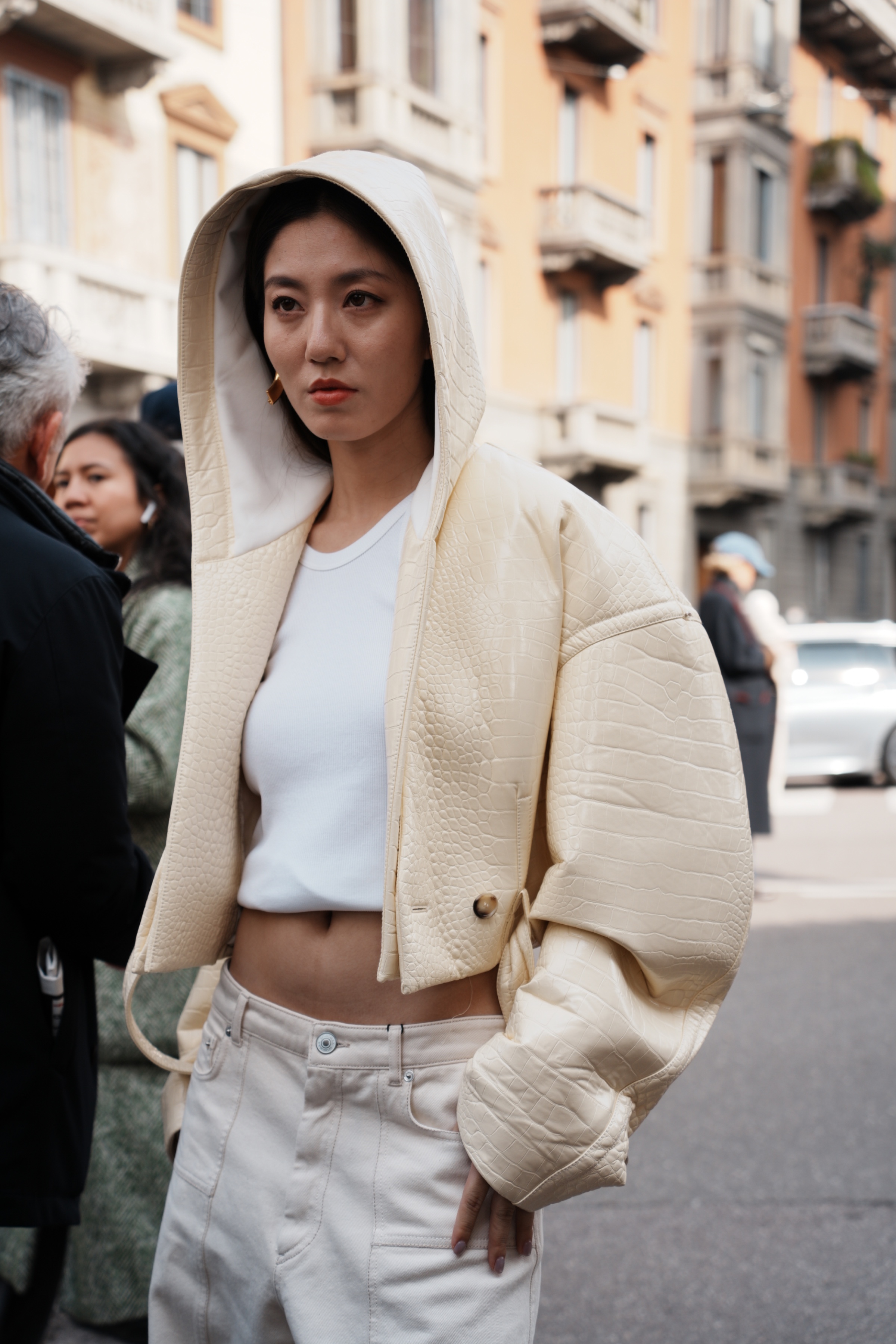 Woman wearing a cream quilted hooded jacket over a white cropped top and white jeans, standing on a city street with one hand in her pocket.