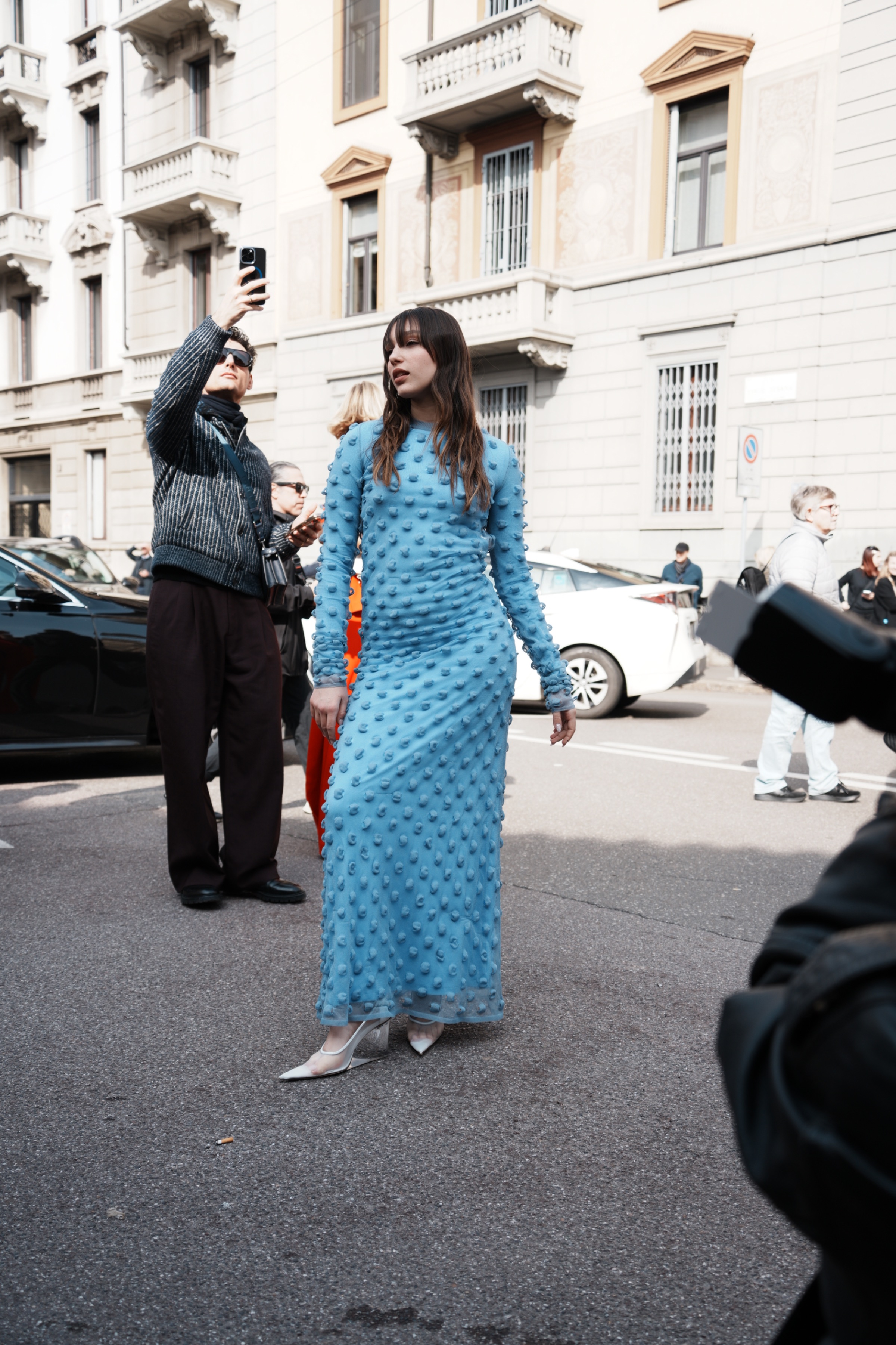 Woman wearing a light blue, sheer long-sleeve dress covered with raised embellishments, paired with white pointed shoes, standing on a city street during the day while people take photos nearby.