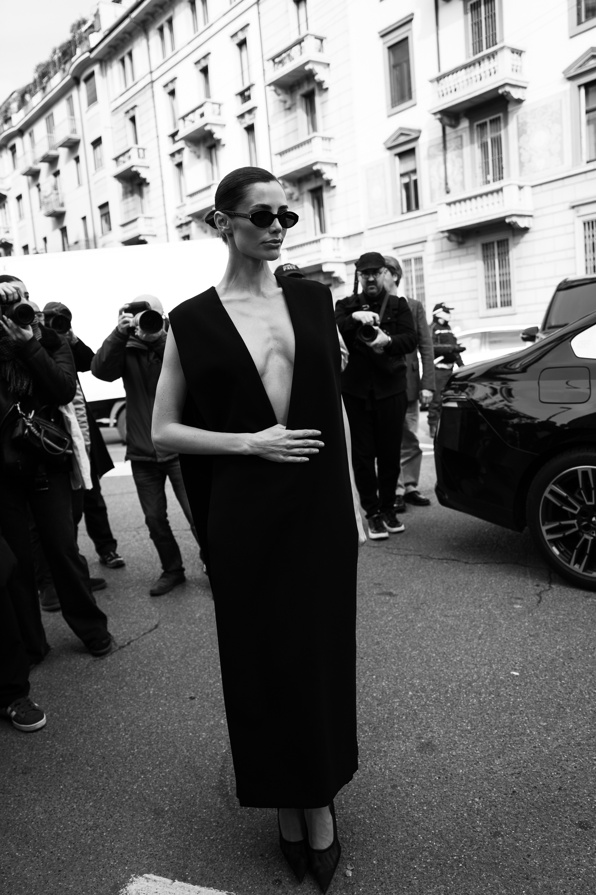 Black-and-white photo of a person wearing dark sunglasses and a sleeveless black dress with a deep V neckline, standing on a city street while photographers take pictures nearby.