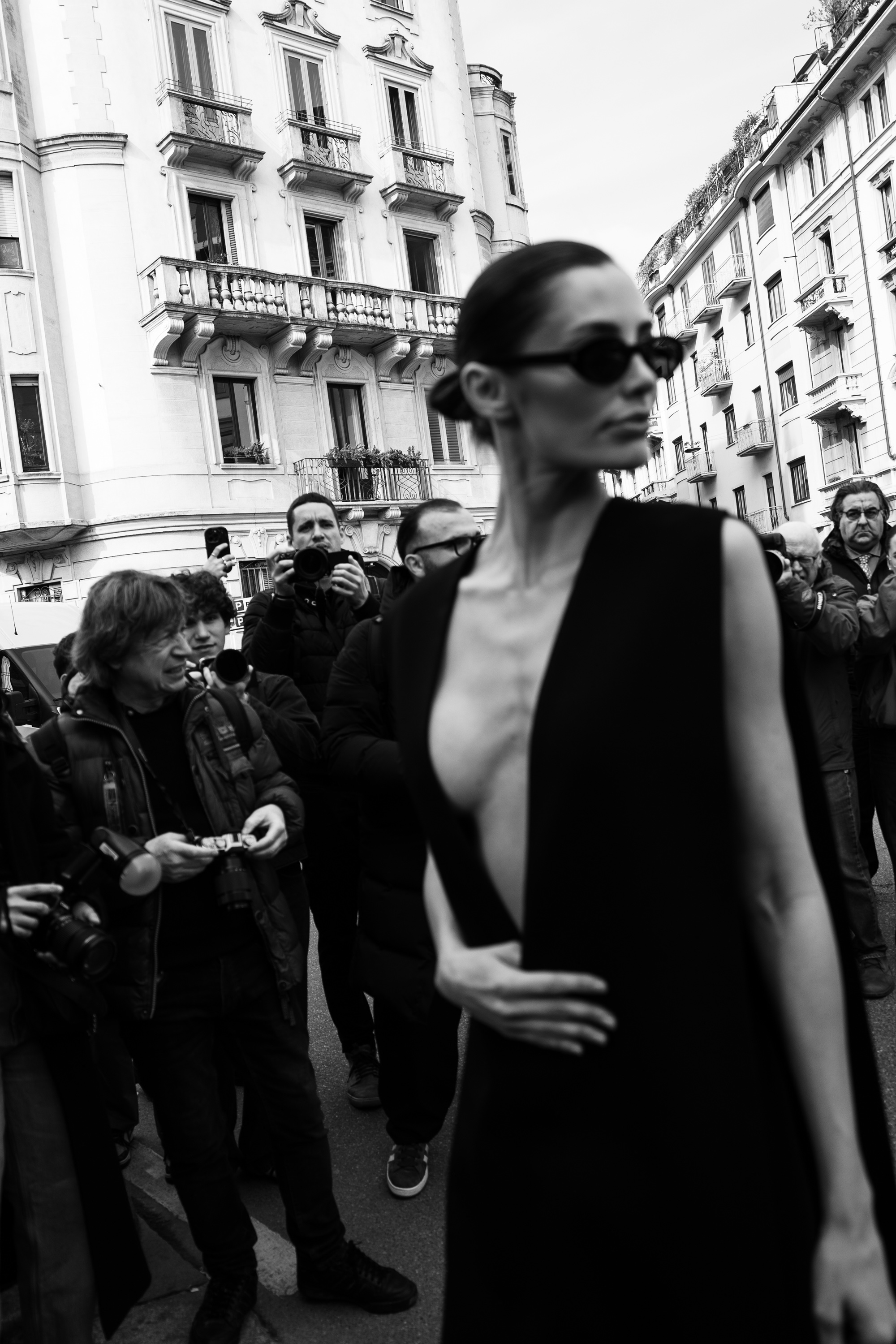 Black-and-white photo of a person wearing dark sunglasses and a sleeveless black dress with a deep V neckline, standing close to the camera as photographers gather on a city street behind them.