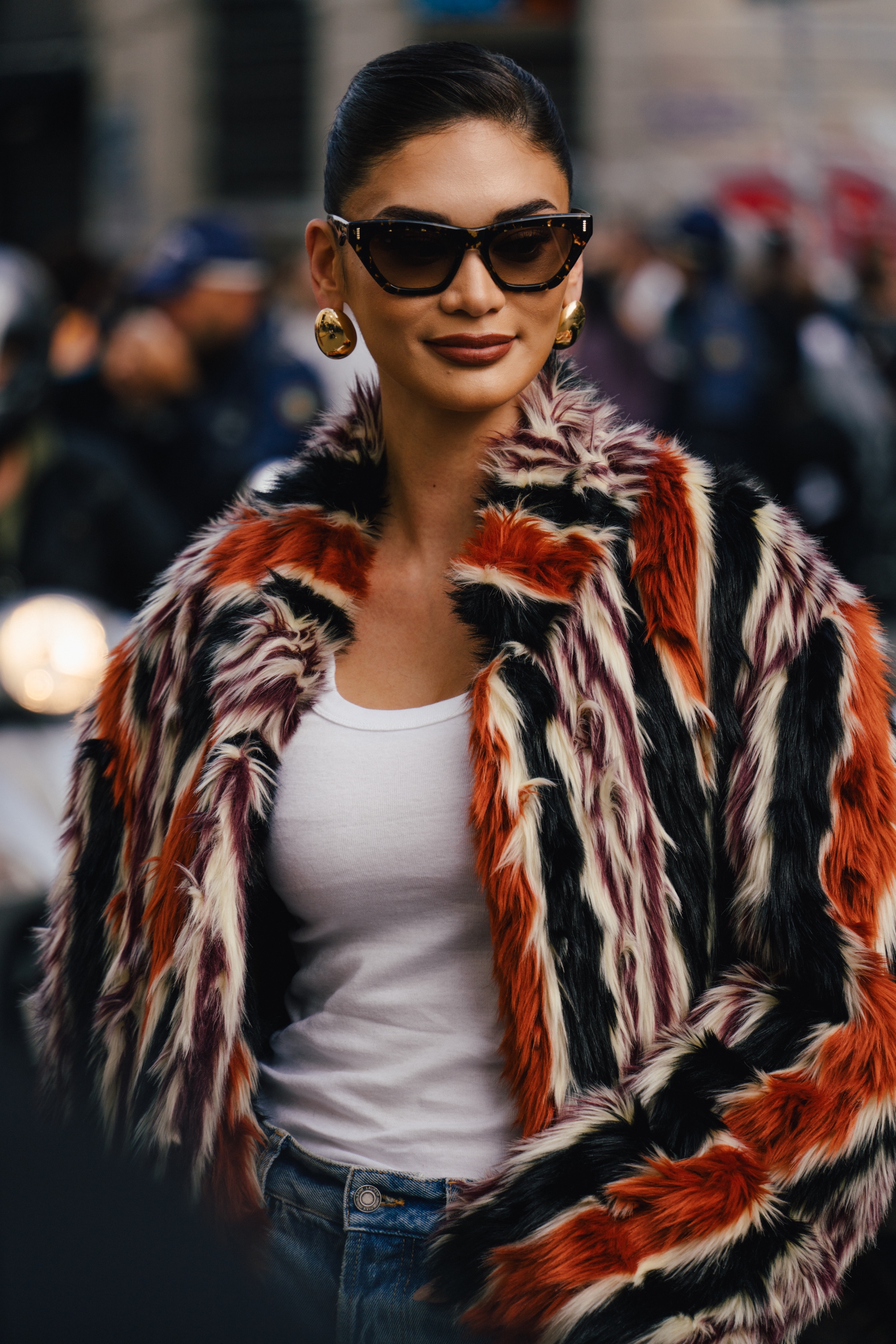 Woman wearing dark cat-eye sunglasses, gold earrings, a colorful striped faux-fur jacket, a white tank top, and blue jeans, standing outdoors with a blurred crowd in the background.