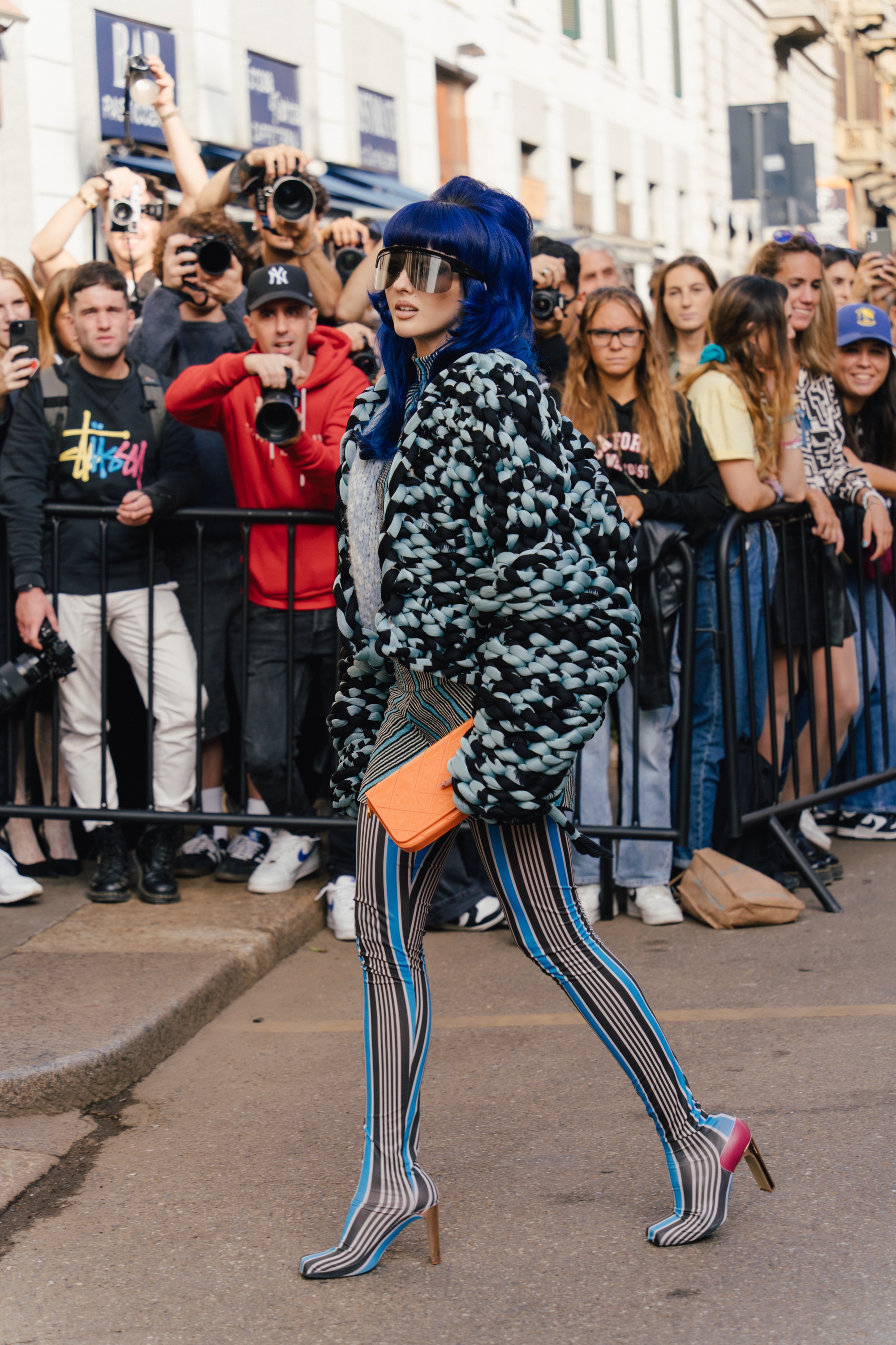 Person with bright blue hair wearing oversized visor-style sunglasses, a chunky textured black-and-mint jacket, striped leggings with pointed heeled boots, and carrying an orange clutch while walking past photographers and onlookers.