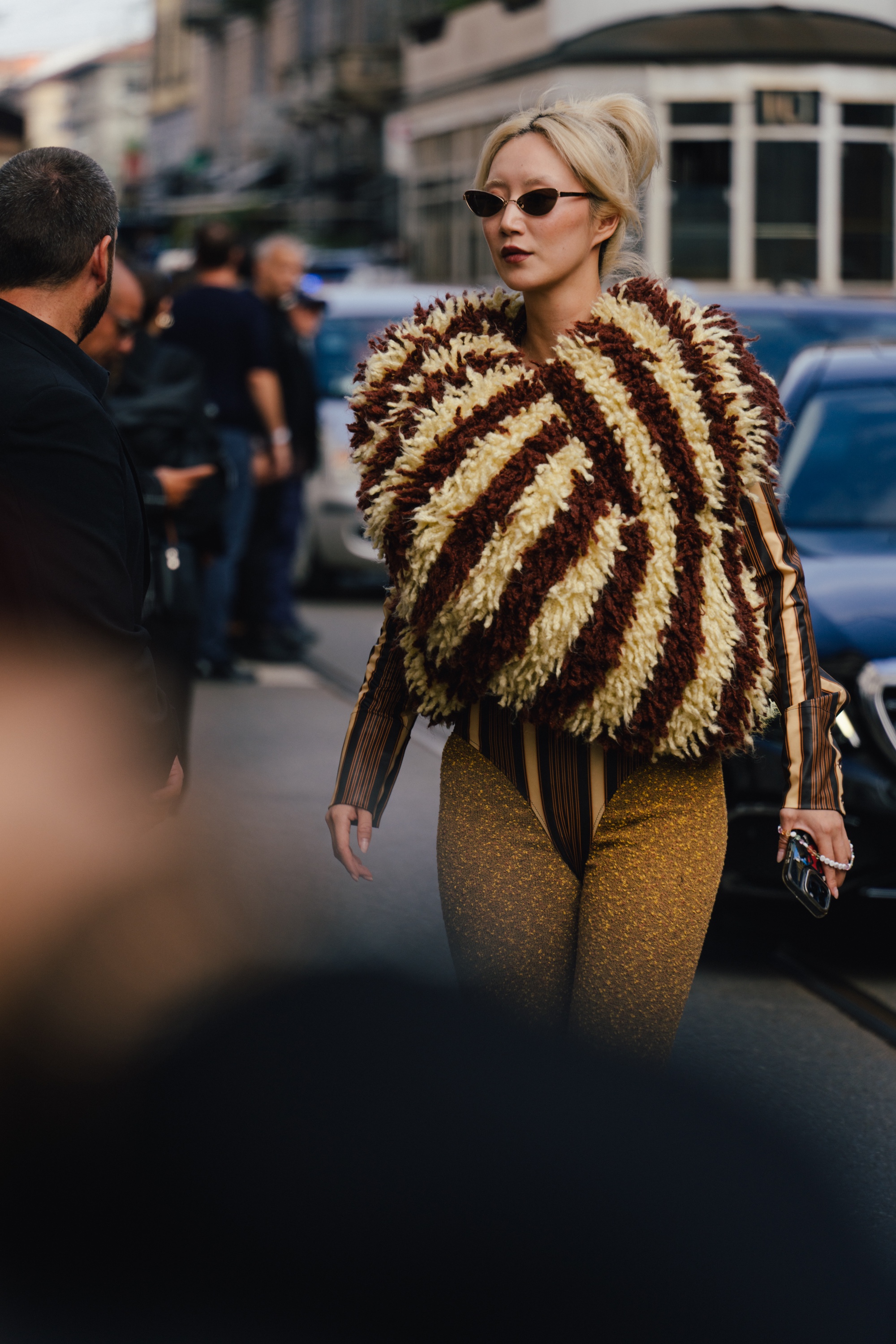 Person wearing narrow black sunglasses, a shaggy cream-and-brown textured top, a striped sheer bodysuit, and glittery gold trousers, walking outdoors while holding a phone.