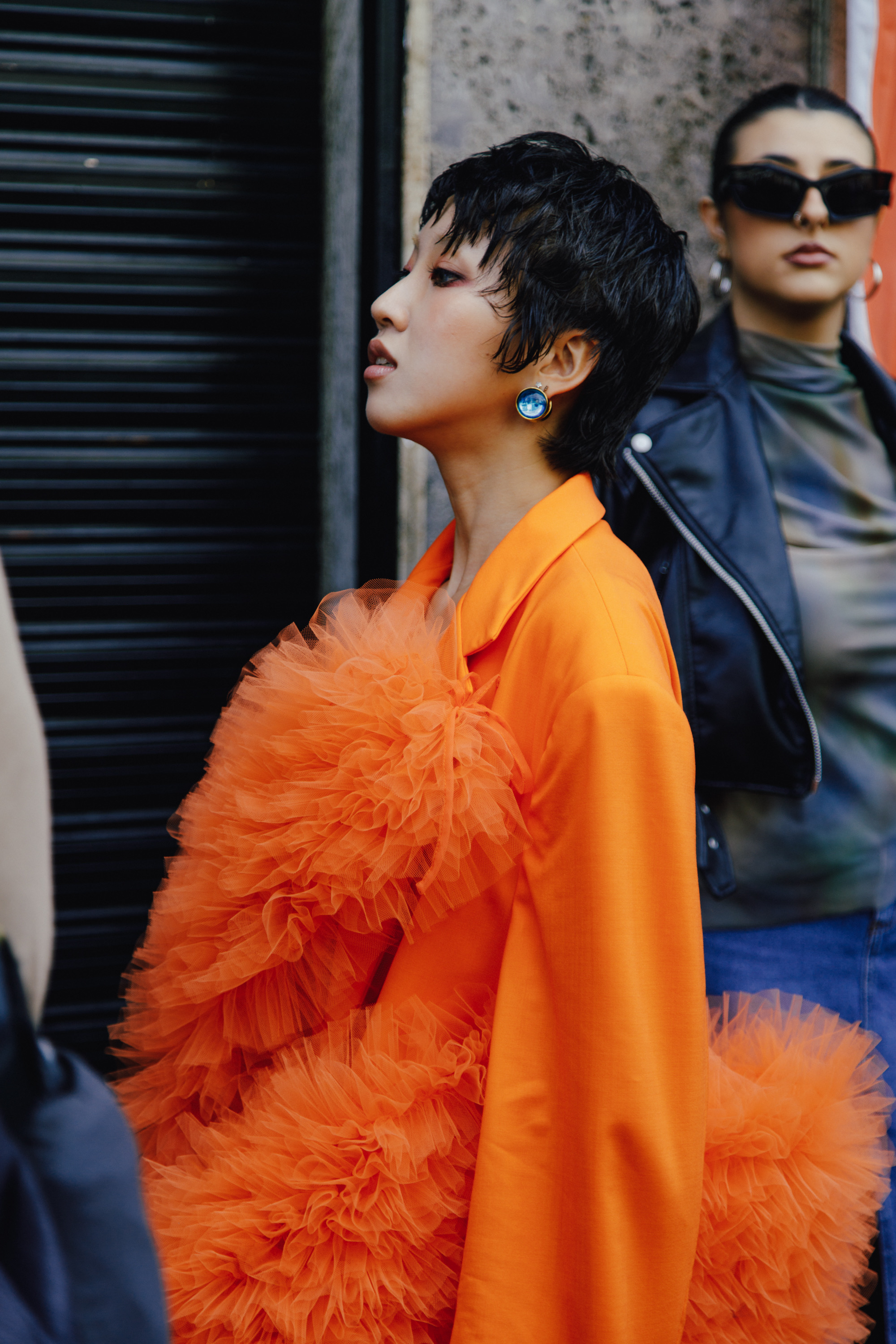 Person with short dark hair wearing a vivid orange outfit with voluminous tulle ruffles, standing in profile beside another person in sunglasses and a black leather jacket.