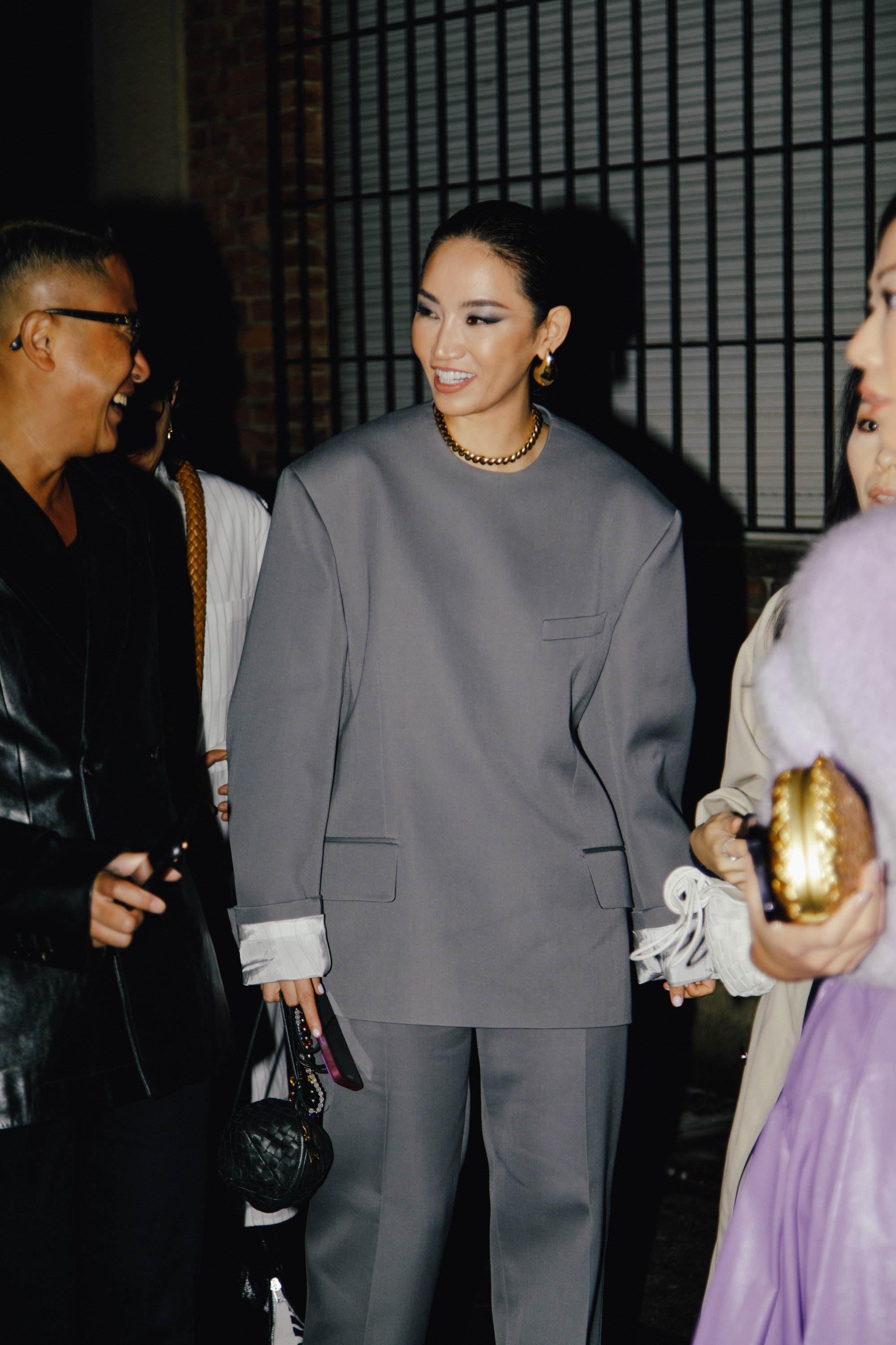 Woman in an oversized gray suit with wide sleeves, wearing gold jewelry and carrying a small black handbag, smiling while standing with other people at night.